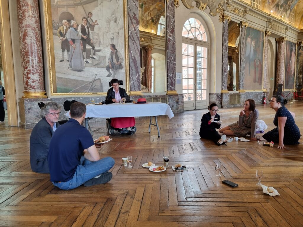 Odile Maurin au centre derrière une table avec une nappe blanche et autour à quelques pas plusieurs conseillères et conseillers municipaux assis par terre faute de siège en train de déjeuner dans la salle des Illustres au Capitole
