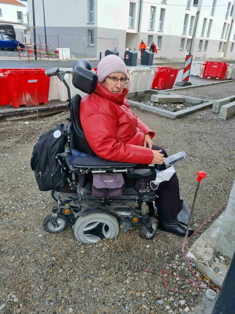 Odile Maurin, coincée rue Avondo, les roues de son fauteuil s'étant enfoncées dans le sol meuble. Elle regarde la caméra avec une moue énervée. 