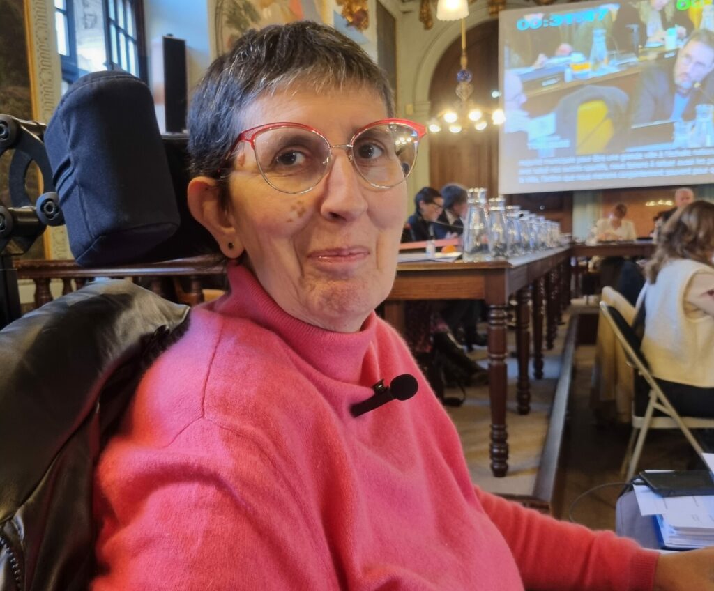 Photo d'Odile Maurin lors du Conseil municipal du 28 novembre 2024 dans la salle du Conseil municipal au Capitole de Toulouse. Odile est vêtue d'un col roulé rose et elle est assise dans son fauteuil roulant électrique.