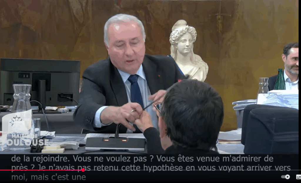 Capture d'écran de la vidéo du Conseil municipal du 26 septembre à Toulouse, où l'on voit le maire Jean-Luc Moudenc arracher le micro de son pupitre des mains de Mme Maurin, qui l'avait simplement tourné vers elle pour lui répondre. Mme Maurin est sur son fauteuil, positionnée en hauteur, devant la tribune. Seule sa tête dépasse de la tribune.