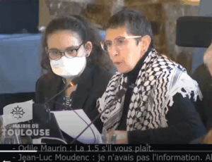 Photo d'Odile Maurin lors du Conseil municipal du 26 septembre 2025 à Toulouse. Elle est assise en train de lire un texte, habillée de noir et portant un keffieh sur les épaules en soutien à la Palestine. Son assistante est à côté d'elle avec un masque FFP2. Odile ne porte pas de masque car elle a vérifié le taux d'aération de la salle.
