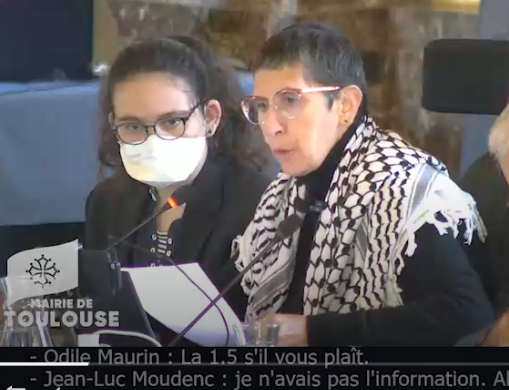 Photo d'Odile Maurin lors du Conseil municipal du 26 septembre 2025 à Toulouse. Elle est assise en train de lire un texte, habillée de noir et portant un keffieh sur les épaules en soutien à la Palestine. Son assistante est à côté d'elle avec un masque FFP2. Odile ne porte pas de masque car elle a vérifié le taux d'aération de la salle.