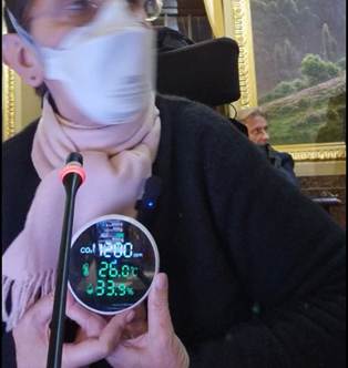 Extrait du direct sur Facebook d'Odile Maurin le 26 novembre 2025 dans la salle du Conseil municipal de Toulouse. Odile porte un masque FFP2 et elle est devant son micro, assise dans son fauteuil roulant, et elle tient entre les mains un capteur de CO2 qui indique 1200 ppm, une température de 26°C et un taux d'humidité de 33,9 %.