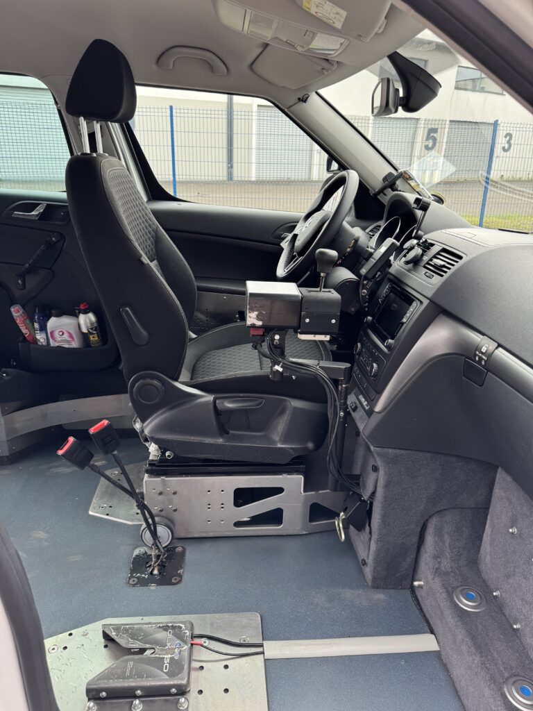 Skoda Yéti blanc de 2017 aménagé conduite en fauteuil avec joystick par ACA à vendre, photo du poste de conduite avec le volant permettant, quand on remet le siège, de conduire normalement et avec le pad à gauche permettant de passer les vitesses, et le frein à main et à droite le joystick. Configuration avec le siège conducteur amovible