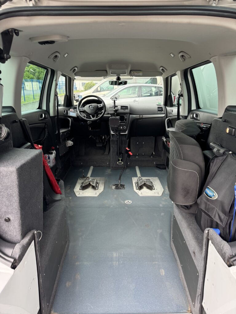 Skoda Yéti blanc de 2017 aménagé conduite en fauteuil avec joystick par ACA à vendre, photo Du poste de conduite avec le volant permettant, quand on remet le siège, de conduire normalement et avec le pad à gauche permettant de passer les vitesses, et le frein à main et à droite le joystick. Et au sol, les ancrages pour deux fauteuils roulants à gauche et à droite ou pour refixer le siège conducteur classique et le siège passager (sièges fournis). À droite, on voit le siège arrière replié qui peut être remis en place, et le véhicule dispose du même siège à gauche qui peut être remonté. Et l'on voit le cheminement pour courir avec les fauteuils roulants pour rejoindre le poste de conduite ou la place passager.