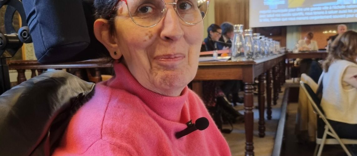 Photo d'Odile Maurin lors du Conseil municipal du 28 novembre 2024 dans la salle du Conseil municipal au Capitole de Toulouse. Odile est vêtue d'un col roulé rose et elle est assise dans son fauteuil roulant électrique.