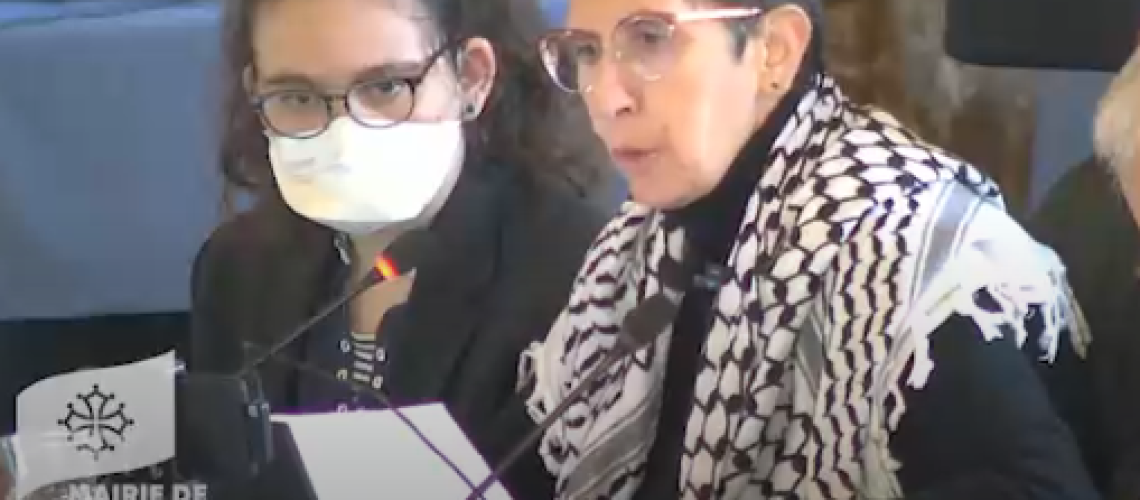 Photo d'Odile Maurin lors du Conseil municipal du 26 septembre 2025 à Toulouse. Elle est assise en train de lire un texte, habillée de noir et portant un keffieh sur les épaules en soutien à la Palestine. Son assistante est à côté d'elle avec un masque FFP2. Odile ne porte pas de masque car elle a vérifié le taux d'aération de la salle.